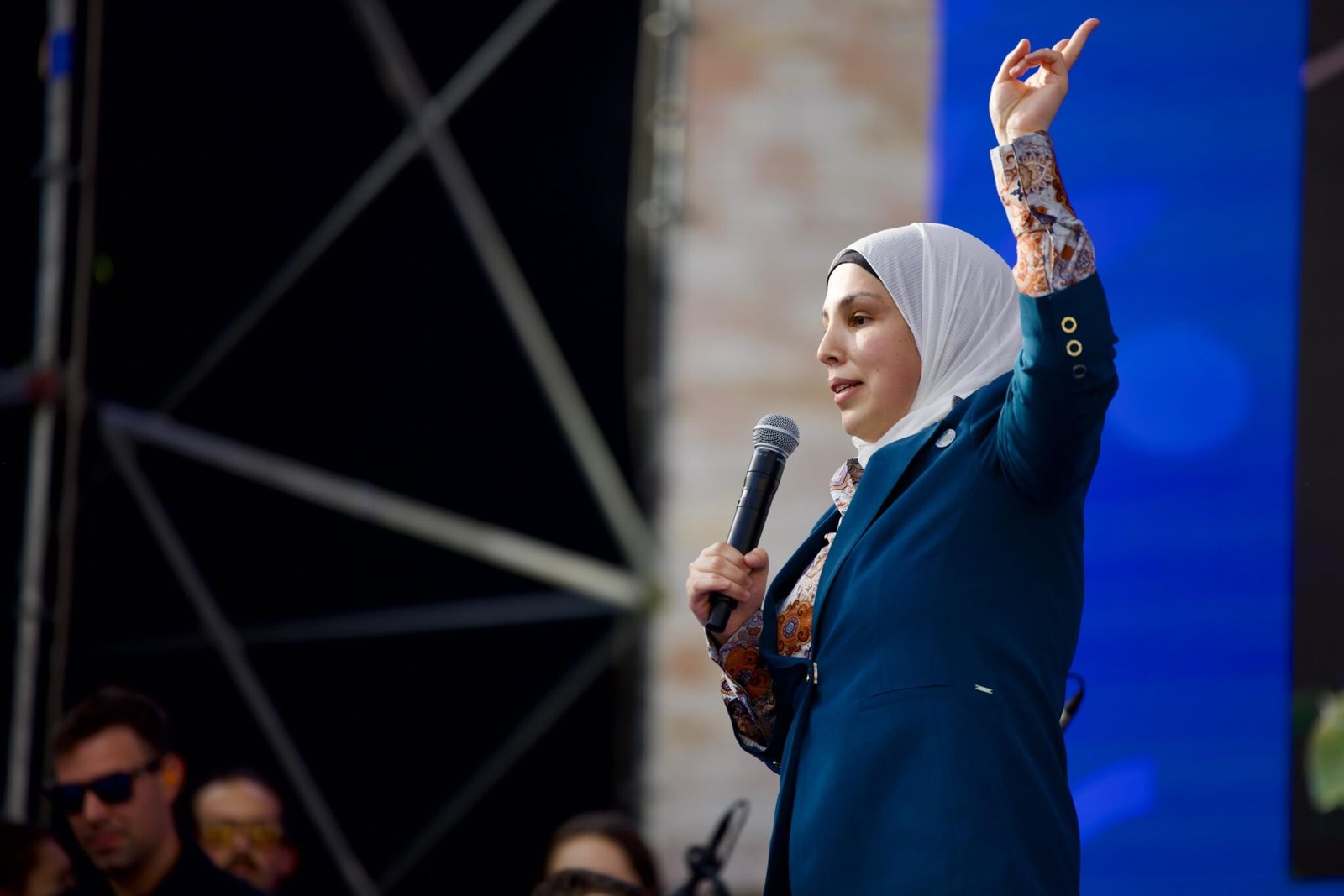 Sara Minkara speaking on stage at the G7, microphone in one hand and the other raised high