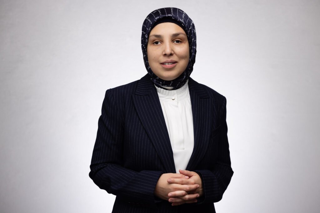 Sara Minkara, smiling, in a navy pinstripe suit and patterned navy hijab against a light gray background