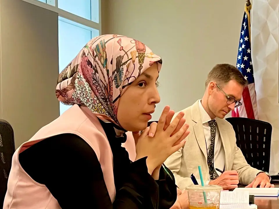 Sara Minkara listening intently in a State Department meeting, hands clasped, with a colleague and an American flag behind her